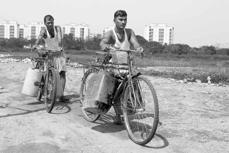 Men transporting drinking water in large containers precariously balanced on bicycles along a dirt road in Mumbai, Maharashtra
